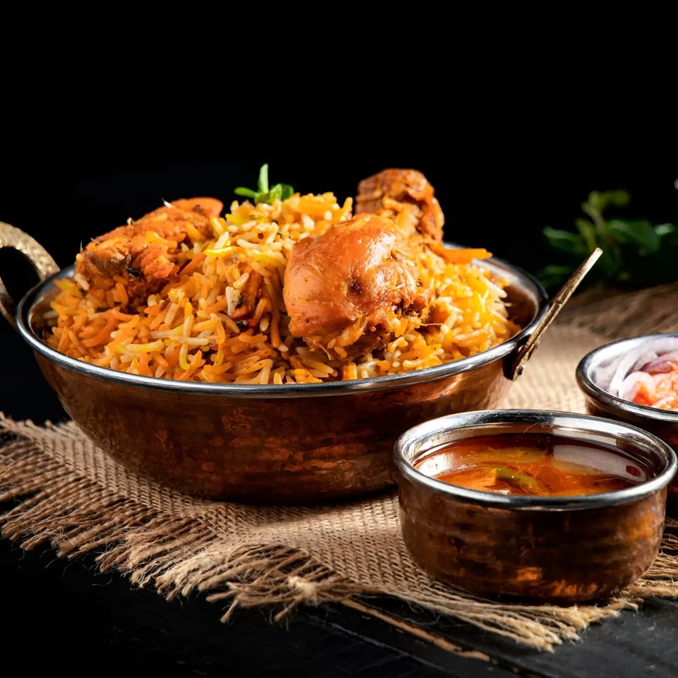 Aachi Biryani's chicken biryani in a copper pot, with two small bowls of sauce and onions.